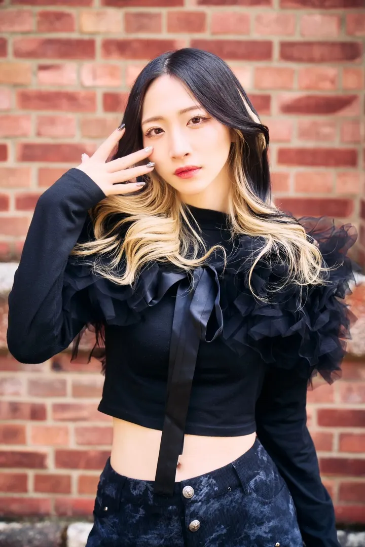 Arisa Sato (佐藤有紗) in a black ruffled top with one hand beside her face, against a brick wall.