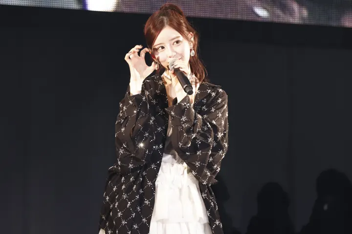 Amane Tsukiashi (月足天音) holds a microphone beside her face, wearing a patterned black jacket over a white tiered dress.