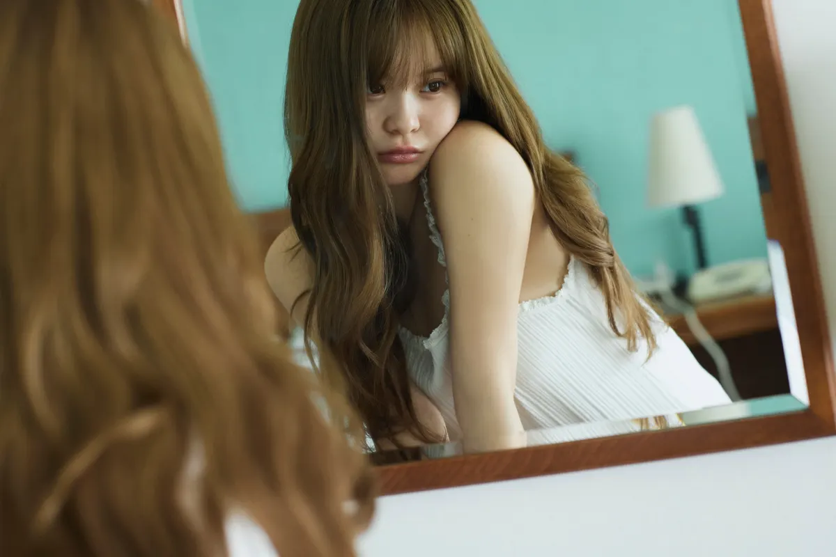 Sakura (さくら)’s reflection in a wooden-framed mirror, wearing a white sleeveless top in a turquoise room.