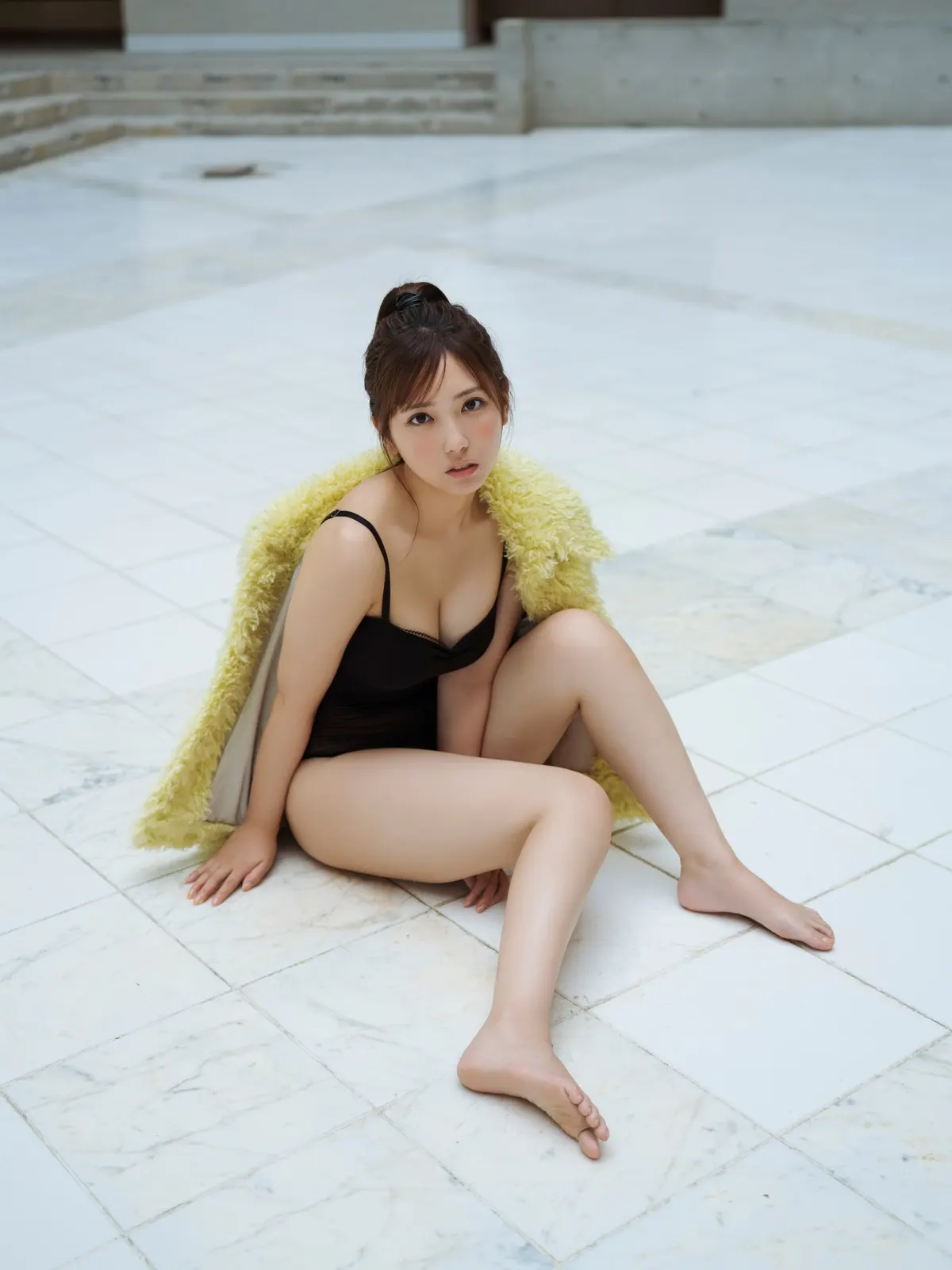 Aika Sawaguchi sits on pale floor tiles in a black outfit with a yellow textured jacket draped around her shoulders for BLTISM VOL.1.
