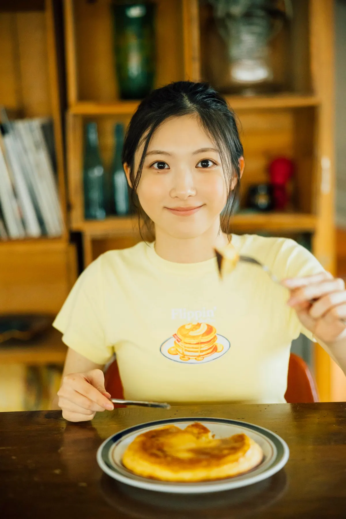 Airi Taniguchi holding cutlery over a pancake at a wooden table, wearing a yellow pancake-print T-shirt.
