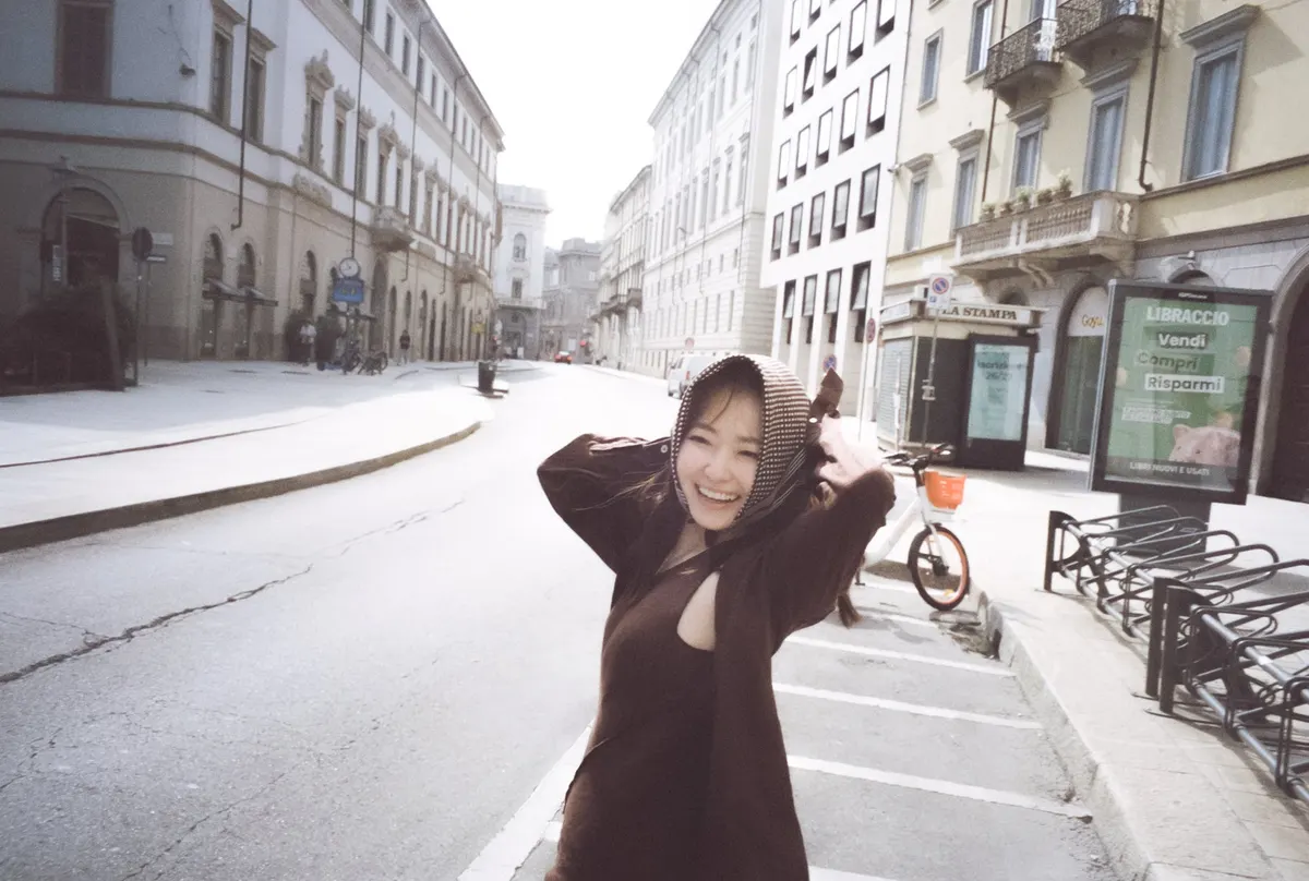 Anna Yamada (山田杏奈) smiles in a dark outfit and patterned headscarf, with both hands beside her head on a street near bicycle racks.