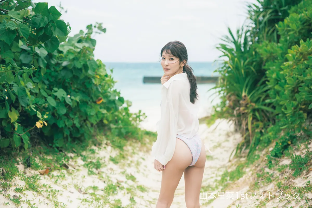 Rina Yamamoto (山本里菜) looking back toward the camera on a sandy path between green plants, wearing a white shirt over patterned swimwear, with the sea in the background.