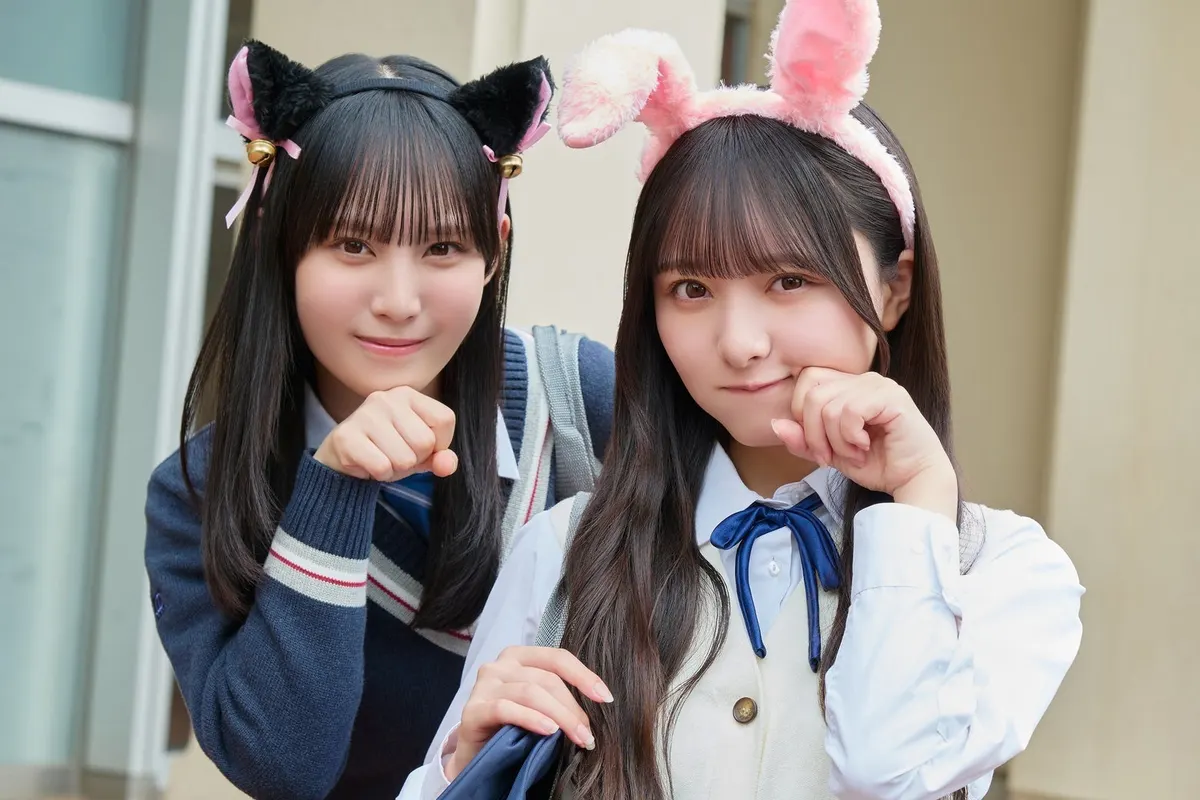 Two Hinakoi campaign performers in school-uniform styling wear black cat-ear and pink rabbit-ear headbands, holding curled hands beside their faces near classroom windows.