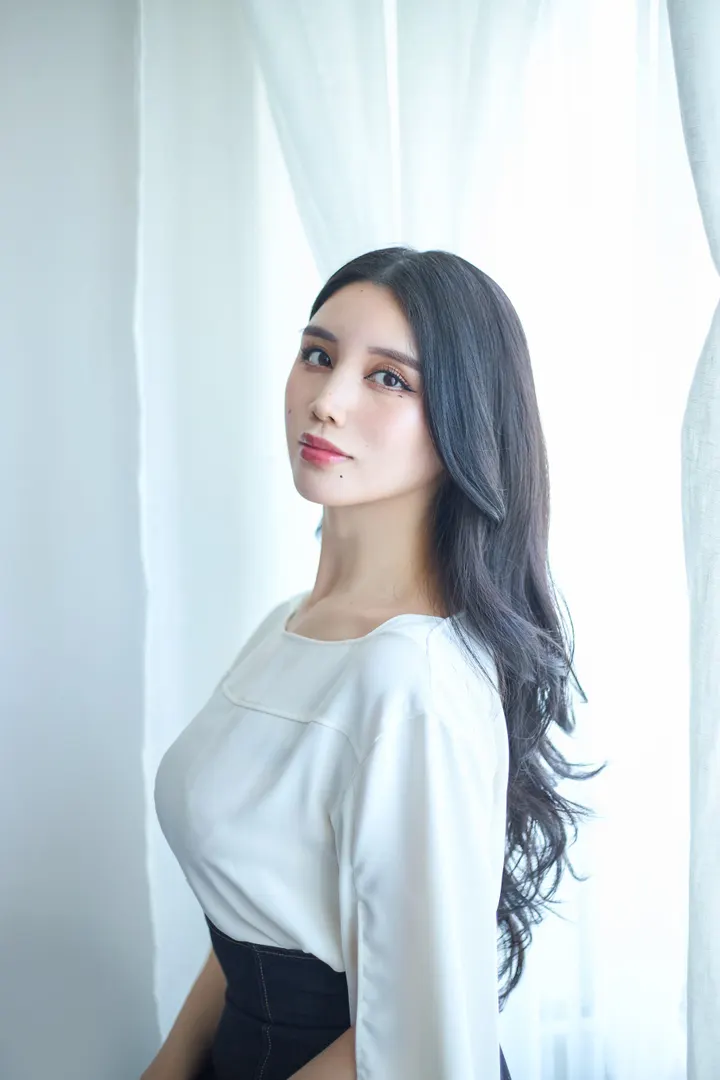 maria (マリア) wearing a white blouse and dark high-waisted garment beside pale curtains.