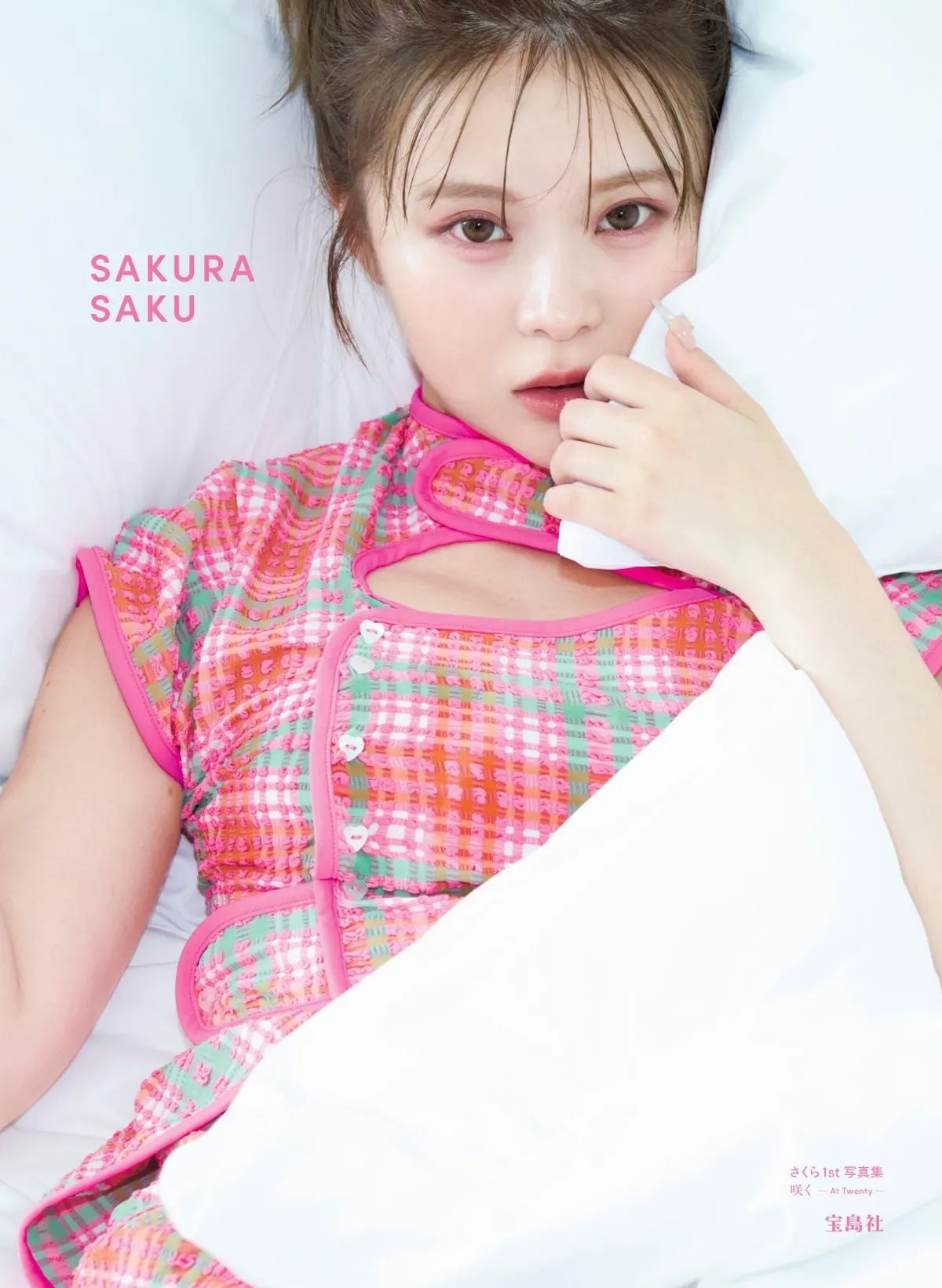 Cover of Sakura (さくら)’s Saku —At Twenty— photobook, showing her in a pink-trimmed checked outfit against white bedding.