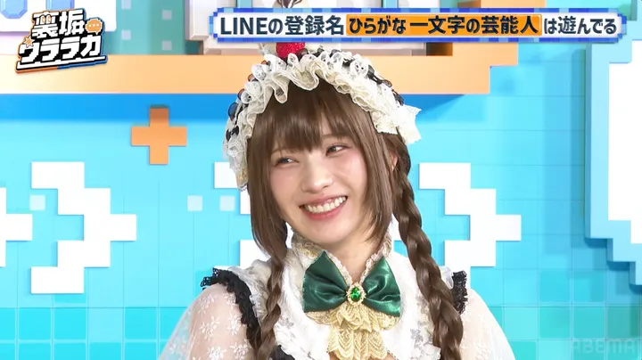 A smiling woman with braided hair wears a lace-trimmed headpiece and a green bow against a turquoise studio backdrop, beneath Japanese program graphics.