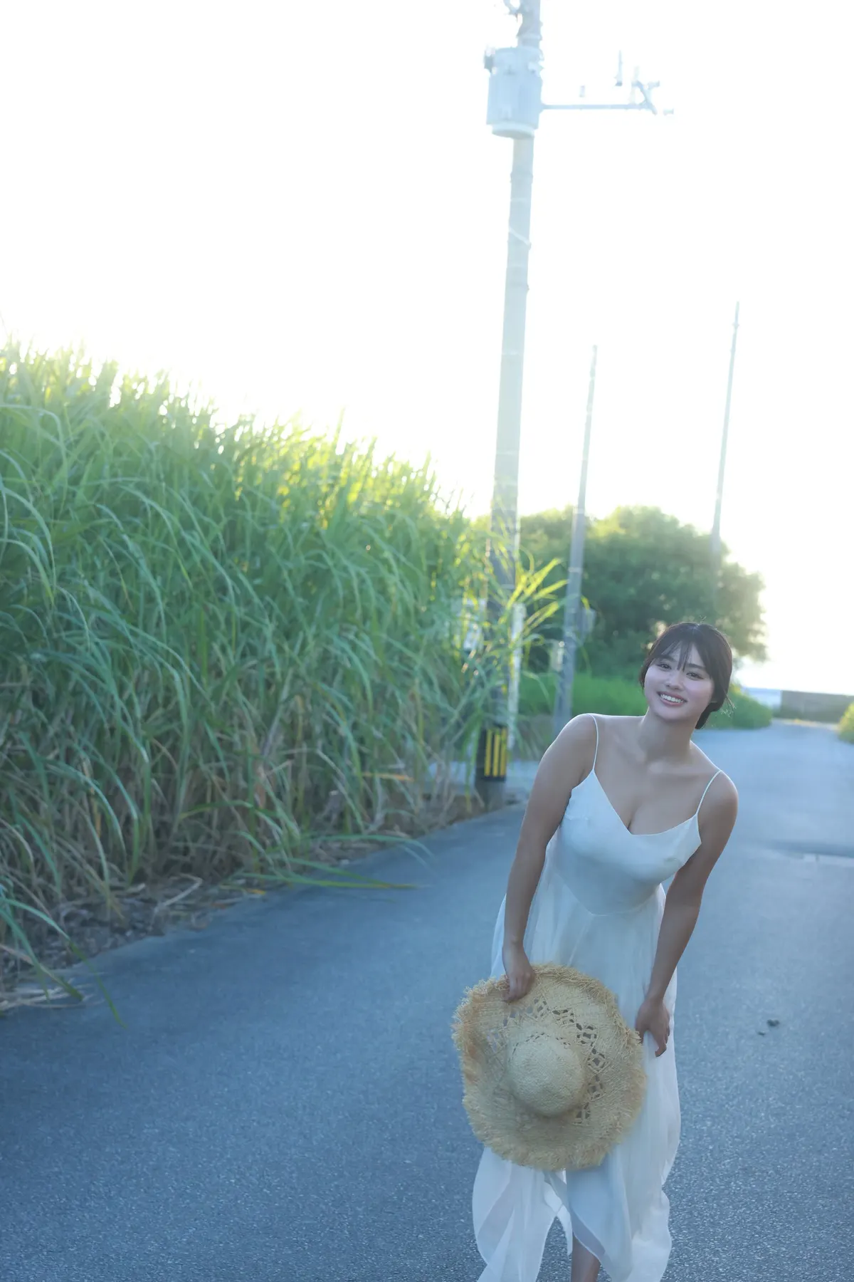 Kokoro Toyoshima (豊島心桜) smiles on a paved road in a white dress, holding a straw hat beside tall grass and utility poles.