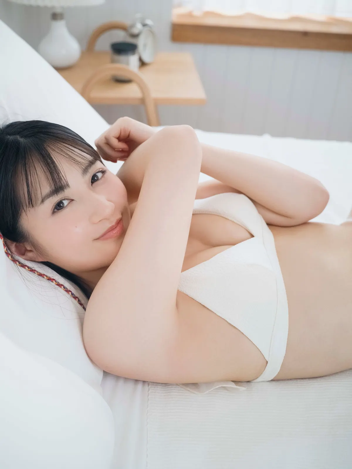 Minami Hyodo (兵頭美波) reclining on white bedding in a white bikini, with a wooden table and chairs behind her.