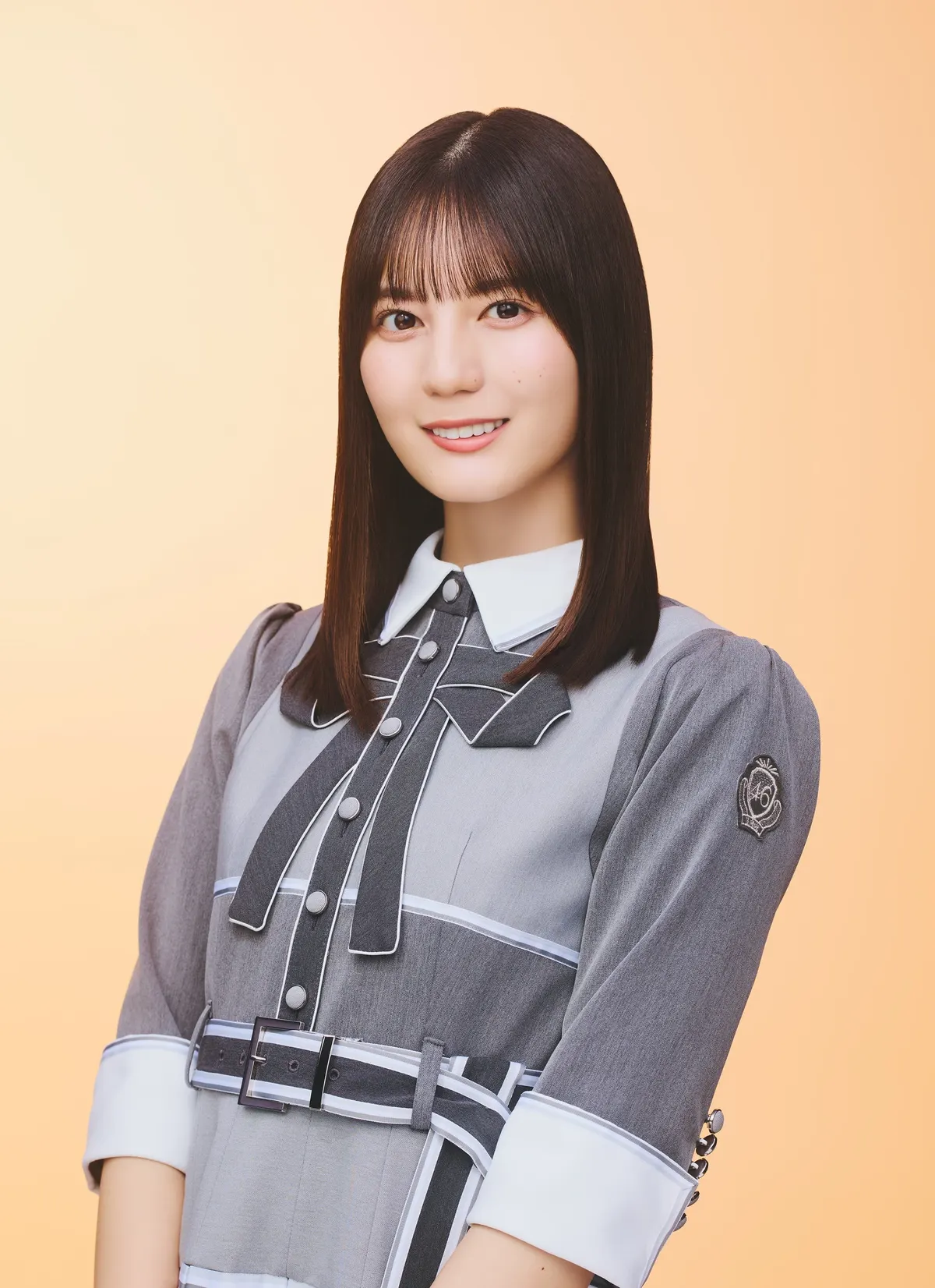 Nao Kosaka wears a gray belted outfit with a white collar and dark ribbon against a pale orange background.