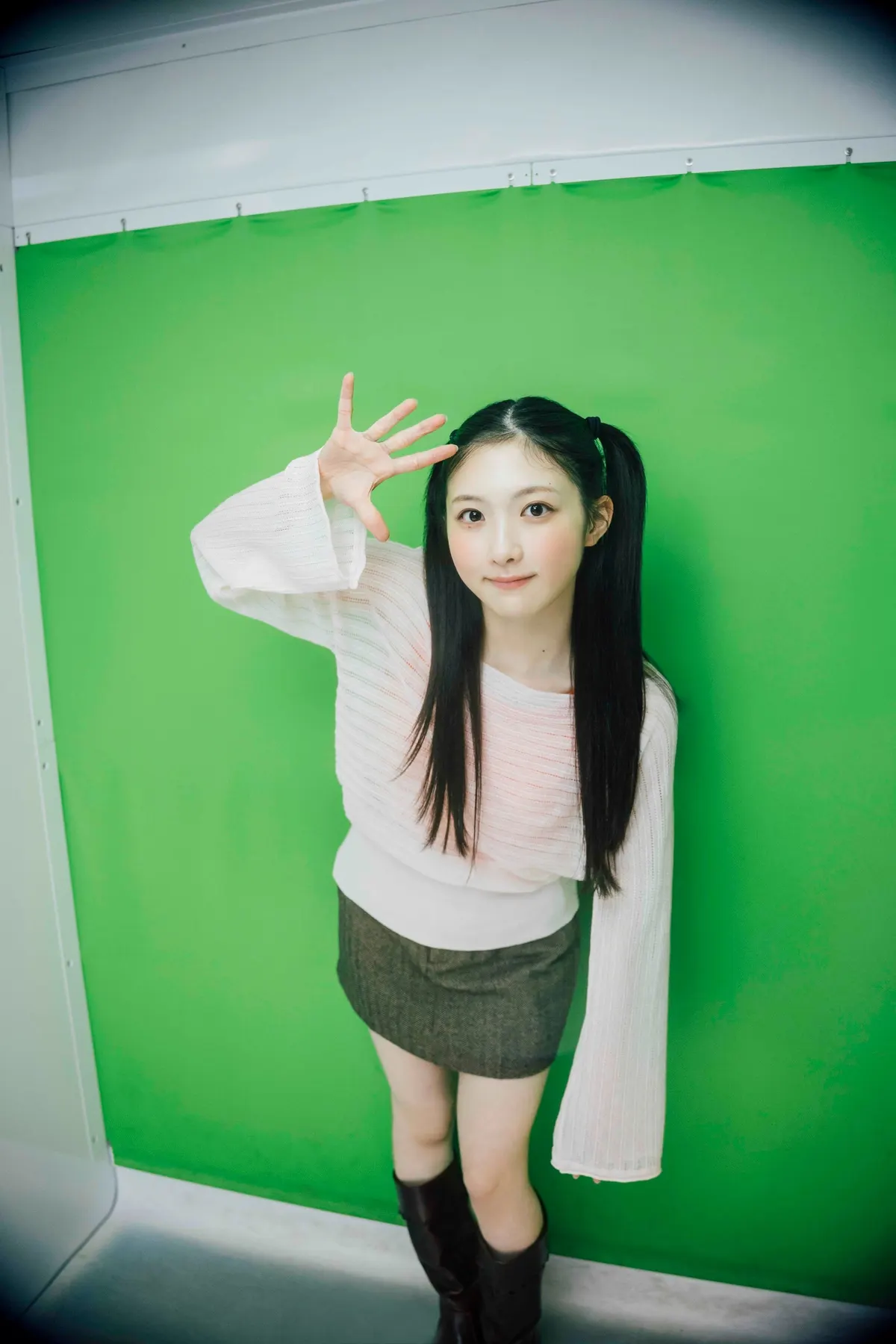 Airi Taniguchi with twin ponytails, a pale long-sleeved top, gray skirt and dark boots against a green backdrop.