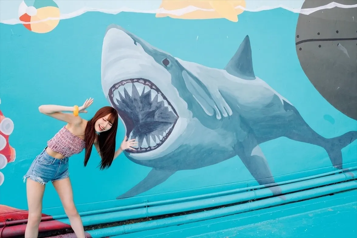 Miyo Nomura (野村実代) posing beside a painted shark mural in a patterned pink top and denim shorts.