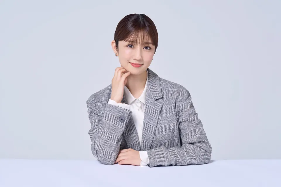 Yuko Ogura (小倉優子) seated behind a pale table in a gray checked blazer and white shirt, with one hand beside her chin.