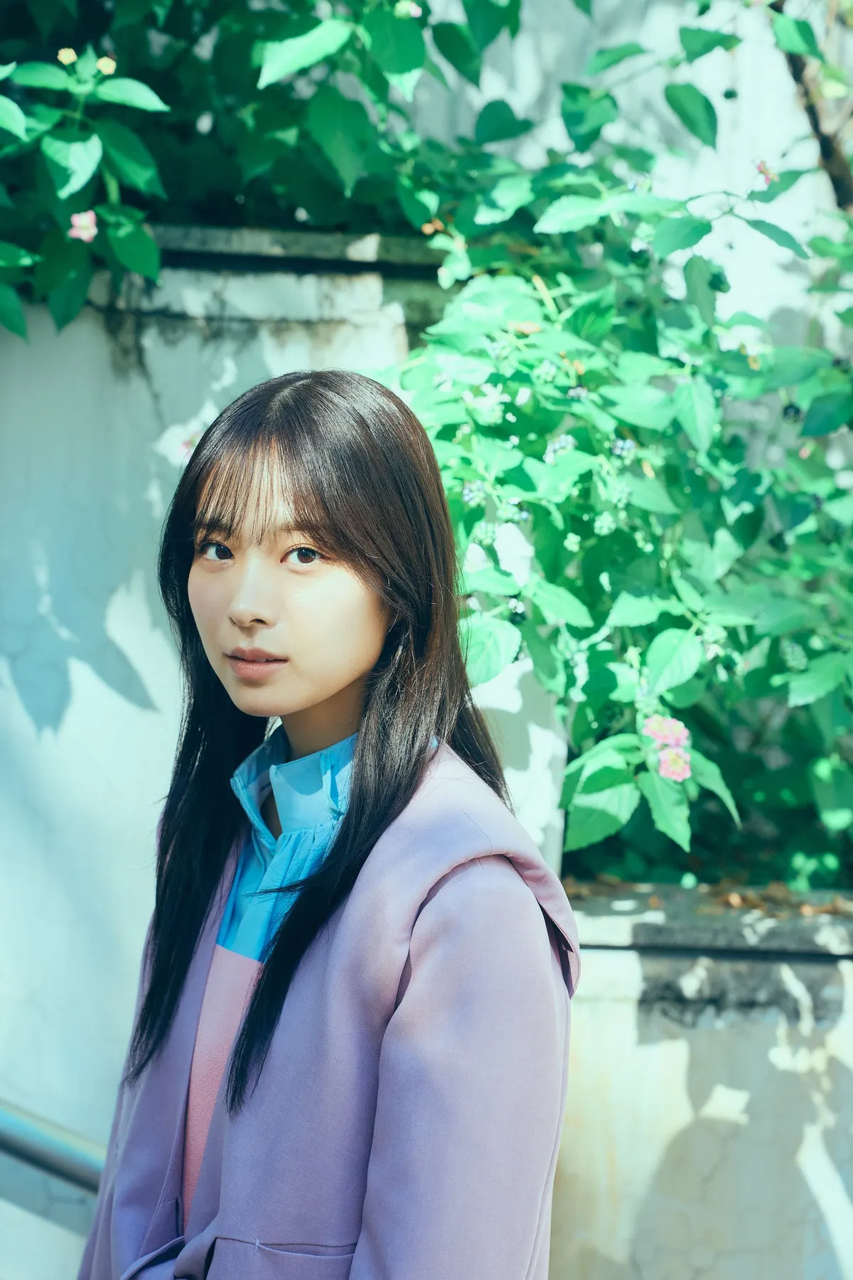 Wako Matsumoto in lavender outerwear with a bright blue collar beside sunlit greenery.