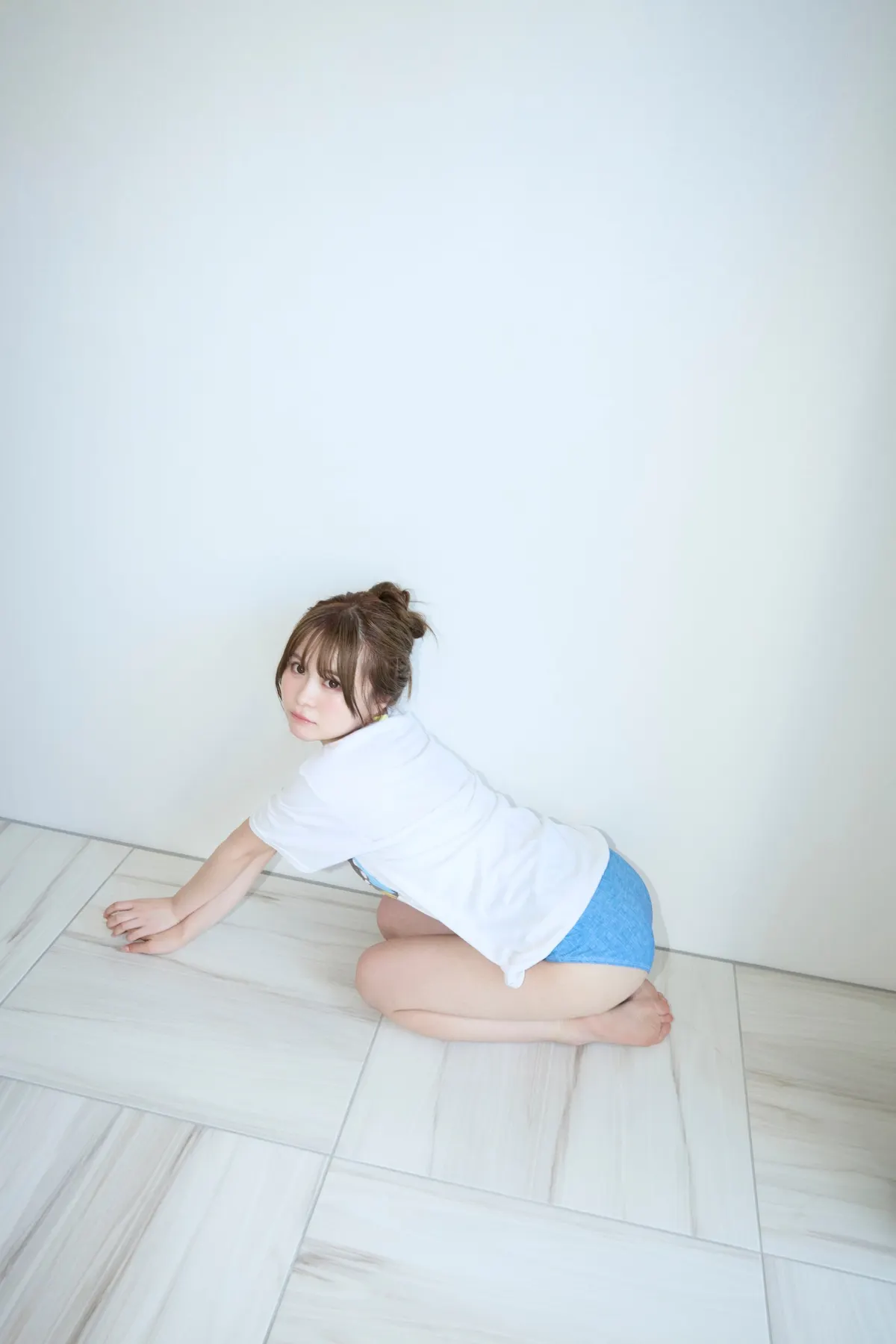 Sakura (さくら) kneeling on pale floor tiles in a white T-shirt and blue bottoms, looking back toward the camera.