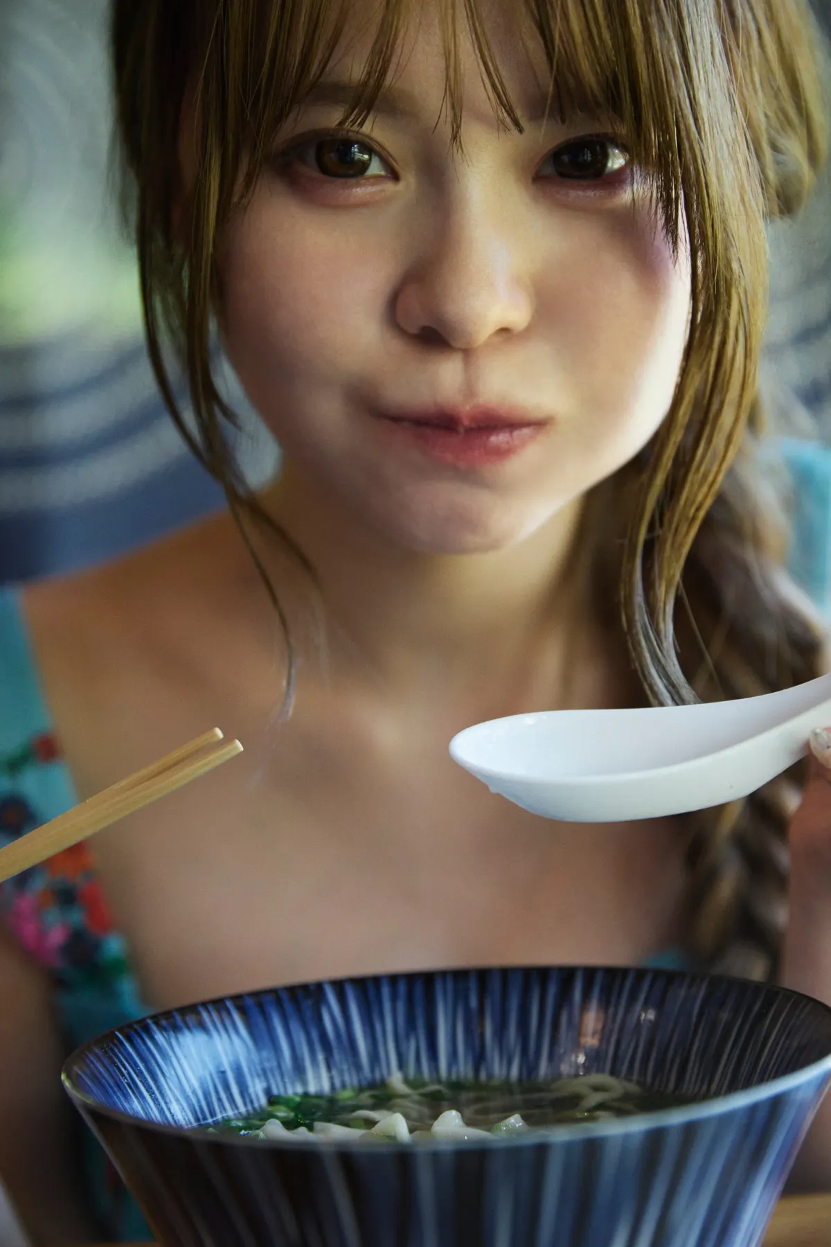 Sakura (さくら) holding a white spoon above a blue bowl of noodles, with chopsticks visible beside her.