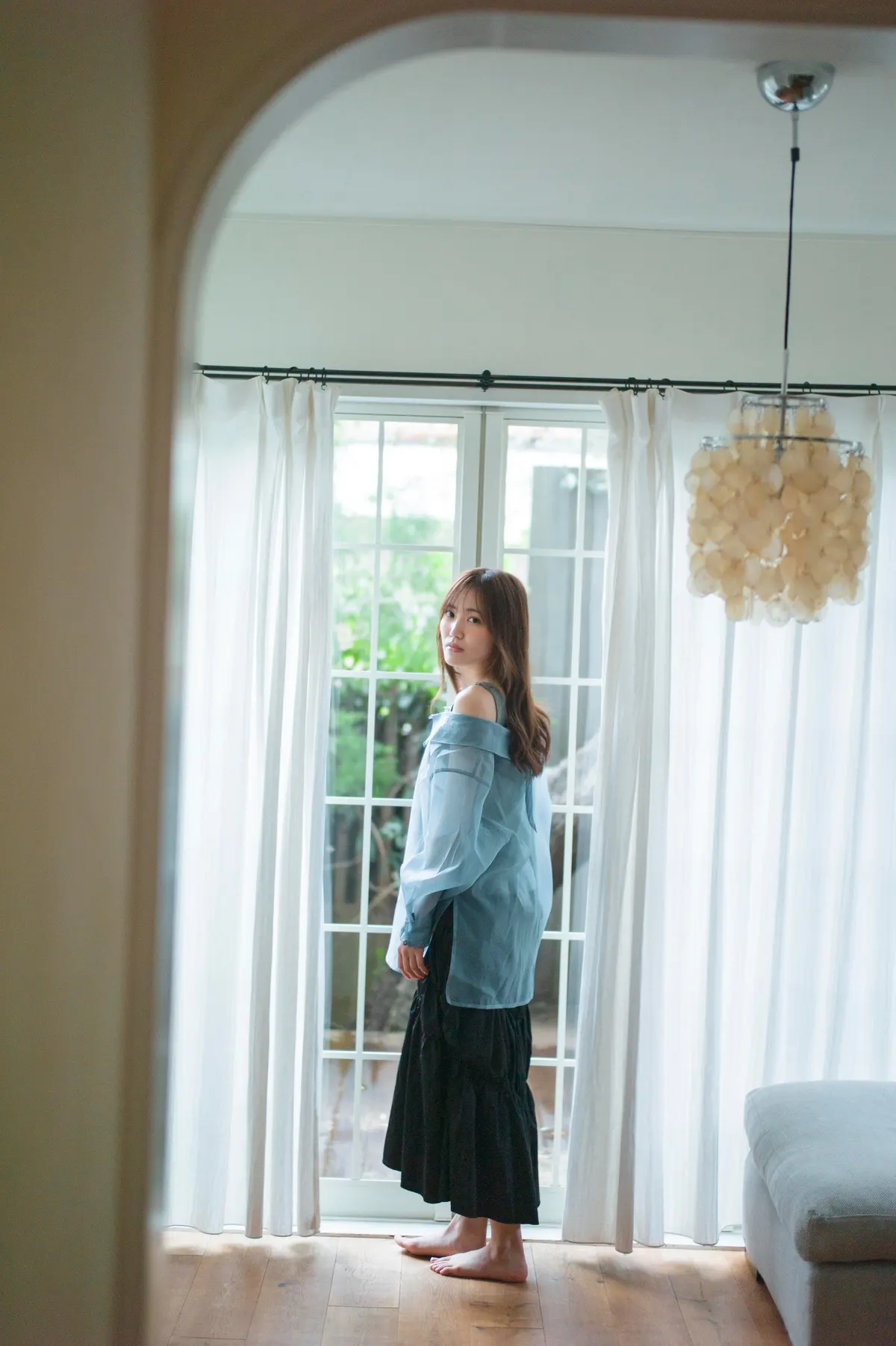 Rina Matsuda standing barefoot beside glazed doors and white curtains in a blue shirt and dark skirt.
