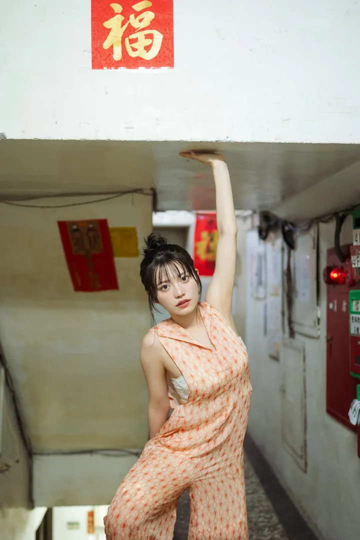Mei Ishihama (石浜芽衣) wearing an orange patterned sleeveless outfit in a stairwell, with one arm raised beneath a low ceiling.