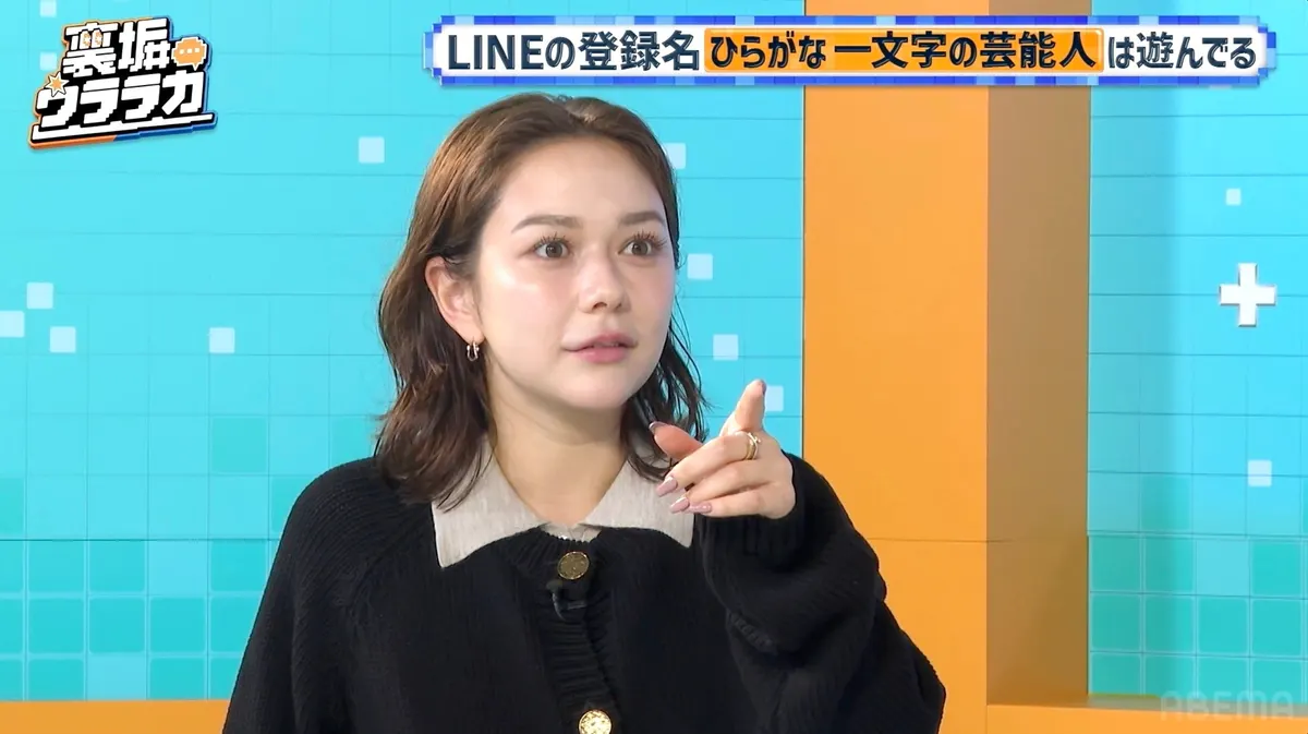 A woman in a black cardigan with a light-colored collar points while seated against a turquoise and orange studio backdrop, beneath Japanese program graphics.