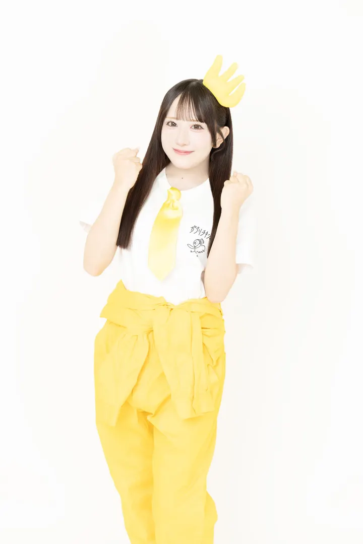 Honoka Aikawa (相川暖花) wearing a white campaign T-shirt, yellow tie, yellow trousers and a yellow head accessory, with both fists raised against a white background.