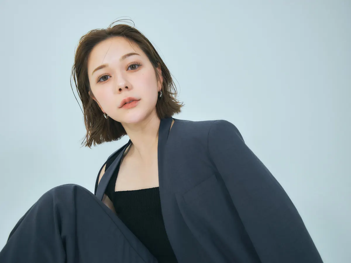 Anna Murashige (村重杏奈) wearing a dark jacket over a black top against a pale blue background.