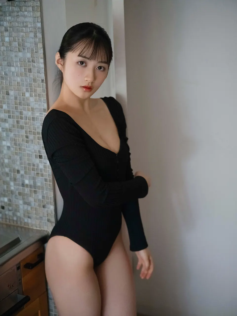 Minami Hyodo (兵頭美波) wearing a black long-sleeved one-piece swimsuit beside a tiled wall and wooden cabinetry.