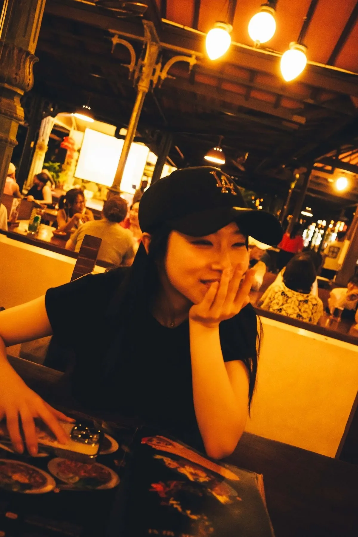 Miyu Kishi smiling with one hand near her face at a restaurant table, wearing a black cap and sleeveless top beneath warm hanging lights.