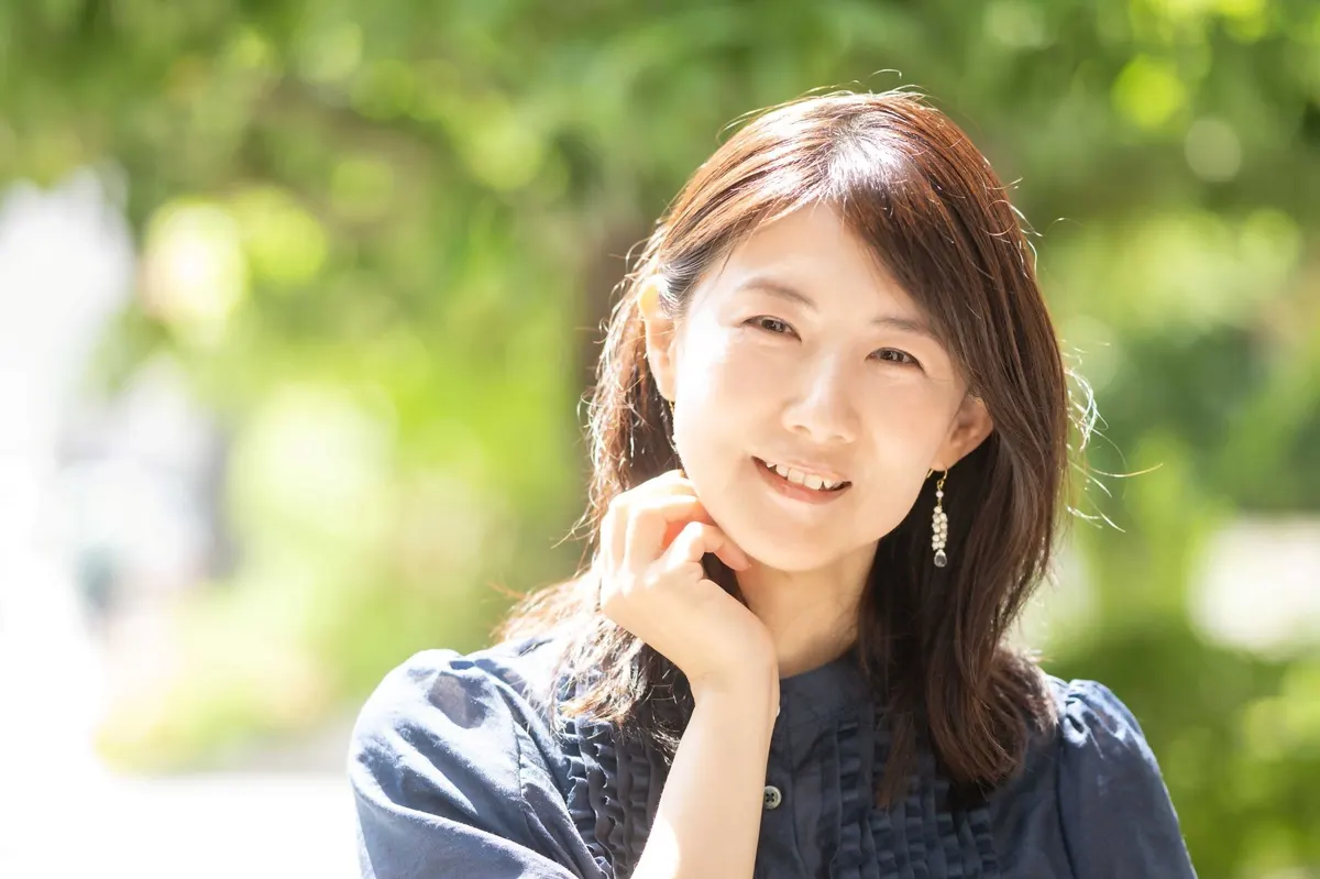Yuri Nakae smiles in a dark blue blouse with one hand beside her face against sunlit greenery.