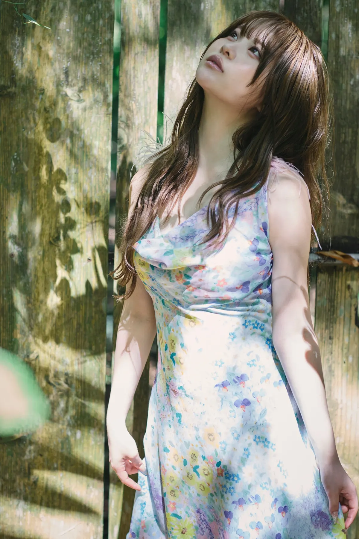 Sakura (さくら) looking upward in a pale floral dress beside weathered wooden boards in dappled sunlight.