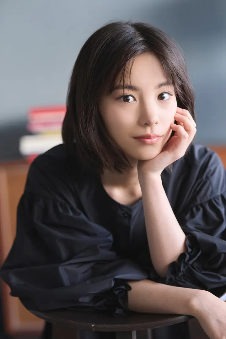 Ami Touma (當真あみ) seated in a dark blouse with one hand resting against her cheek.