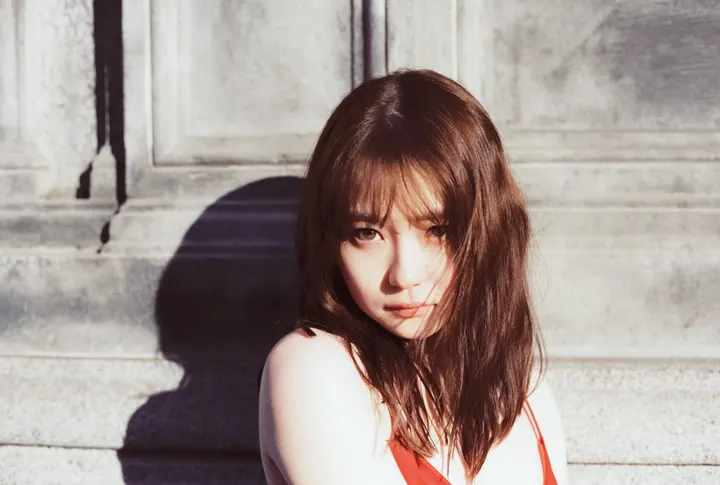 Anna Yamada (山田杏奈) with shoulder-length brown hair and red shoulder straps, lit by direct sunlight against a gray paneled wall.