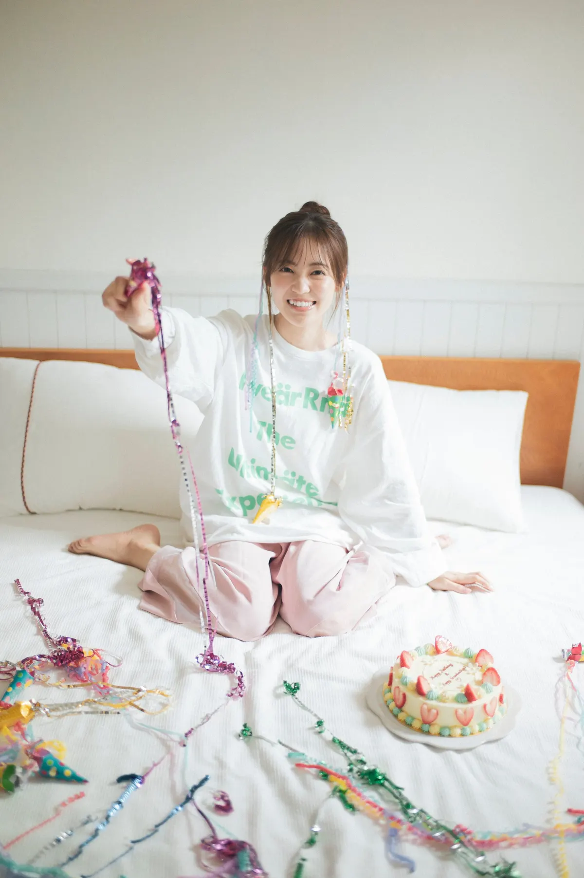 Rina Matsuda seated on white bedding beside a decorated cake, holding colorful party streamers.
