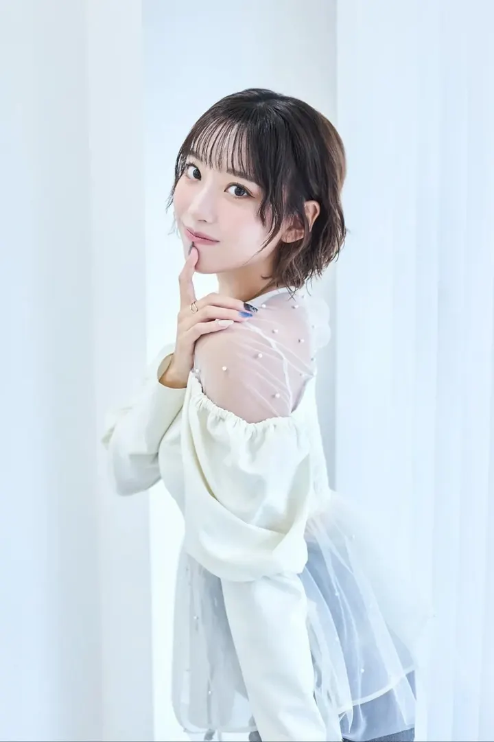 KURUMIWARININGYO (くるみ割り人形【くるみん】) looking back toward the camera with a finger near her chin, wearing a white top with sheer dotted fabric.