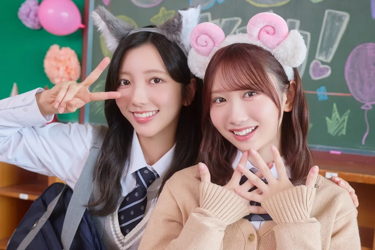 Two Hinakoi campaign performers in school-uniform styling wear pointed animal-ear and curled-horn headbands beside a classroom chalkboard decorated with drawings and balloons.