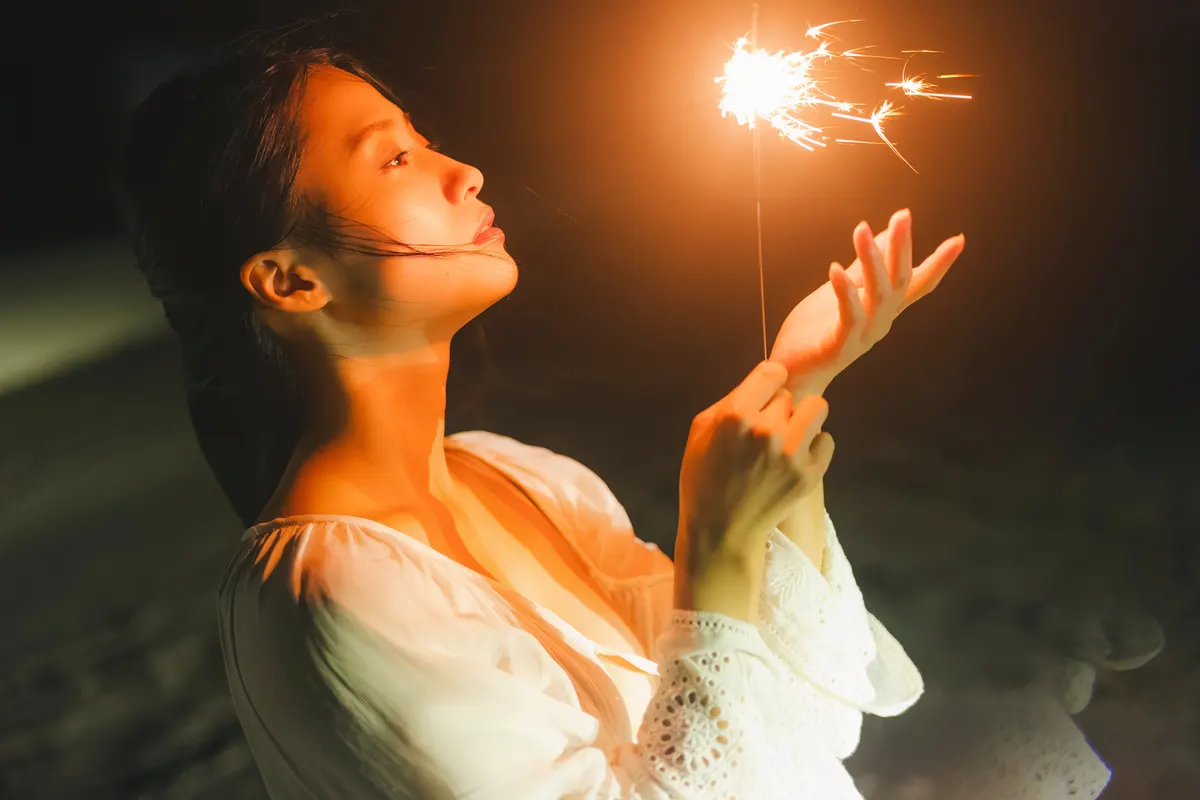 Ayano Sumida watches a glowing sparkler at night while wearing a white blouse with lace cuffs for BLTISM VOL.1.