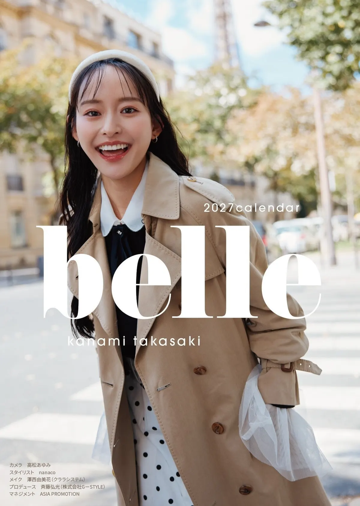 Cover of Kanami Takasaki’s 2027 desk calendar belle, showing her smiling in a beige trench coat and white headband on a tree-lined street.