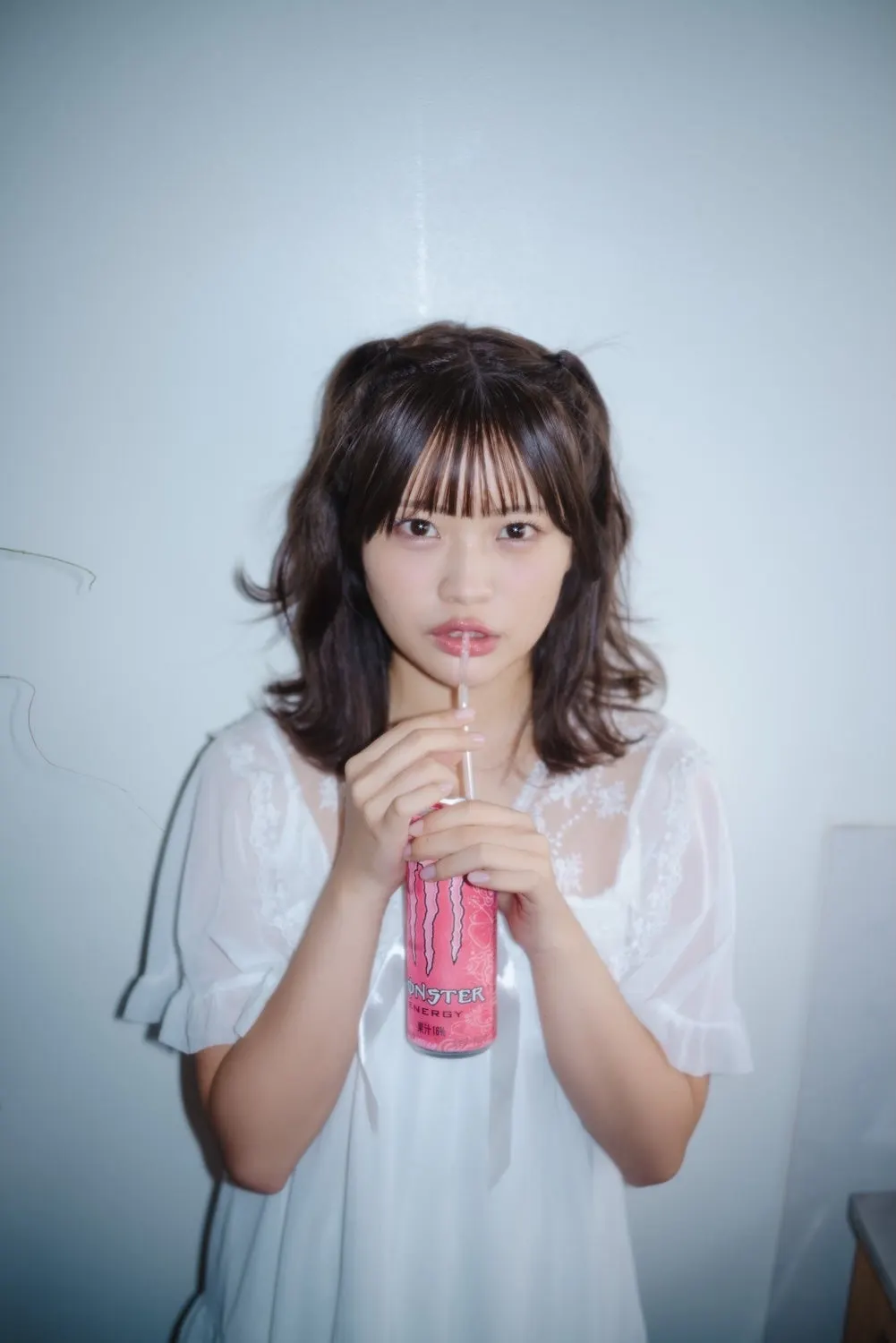 Mii Sugii holding a pink drink can with a straw, wearing a white lace-trimmed garment against a pale wall.