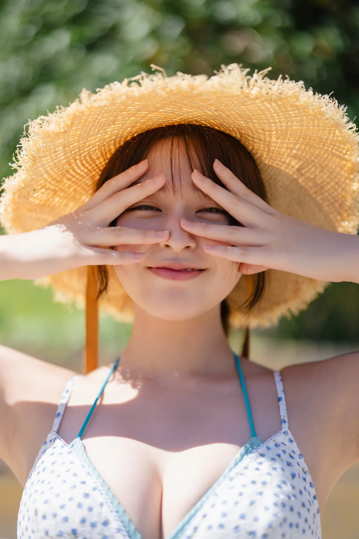 An Yamamoto wearing a broad straw hat and patterned swimwear, framing her eyes with her fingers outdoors in an advance photograph for her first photobook.