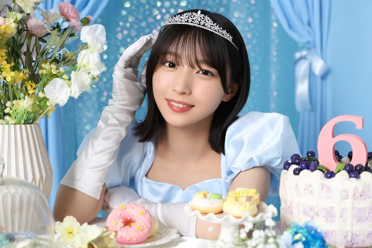 Hinakoi anniversary portrait of a short-haired performer in a pale blue dress and tiara raising a white-gloved hand beside flowers, doughnuts and a cake with a pink number six.