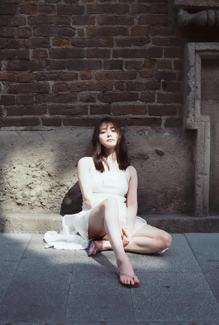 Anna Yamada (山田杏奈) sits on stone pavement in a white sleeveless dress and pale pink sandals against a brick-and-stone wall.
