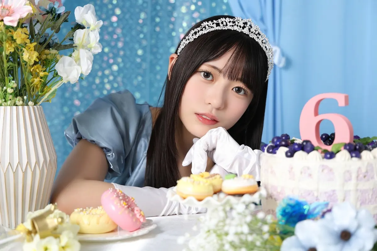 Hinakoi anniversary portrait of a performer in a blue dress and tiara resting her chin on a white-gloved hand beside flowers, doughnuts and a cake topped with a pink number six.