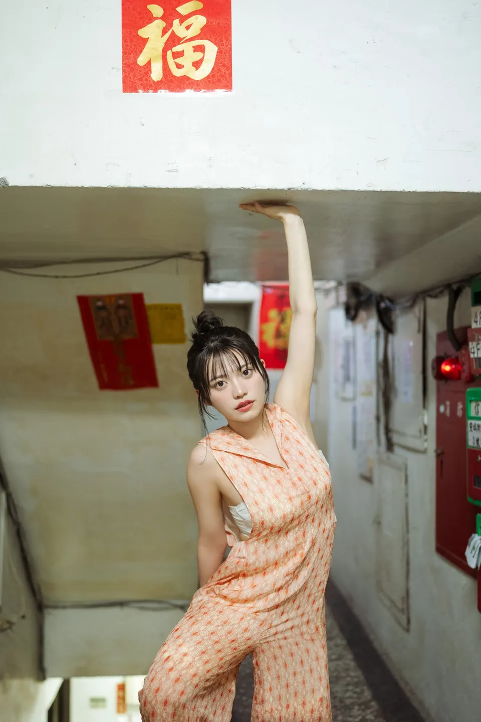 Mei Ishihama (石浜芽衣) wearing an orange patterned sleeveless outfit in a stairwell, with one arm raised beneath a low ceiling.