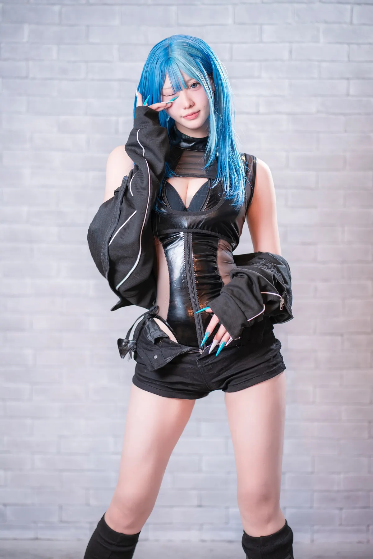 Publicity portrait of ZIRO with blue hair and a black costume, winking with fingers beside one eye against a pale brick wall.