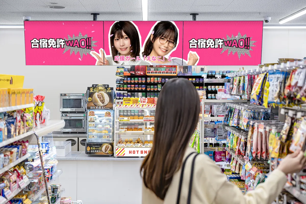 Convenience-store promotional image with Enako (えなこ) and Umi Shinonome (東雲うみ) on overhead pink WAO!! screens and a shopper seen from behind.