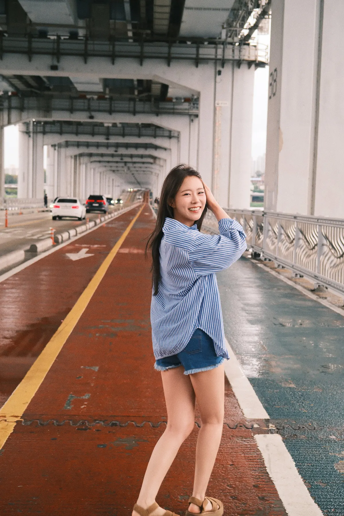 Han Seung Yeon (ハン・スンヨン) smiles on a marked path beneath a bridge, wearing a blue-and-white striped shirt and denim shorts.