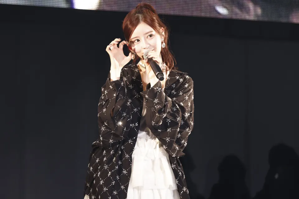Amane Tsukiashi (月足天音) holds a microphone beside her face, wearing a patterned black jacket over a white tiered dress.