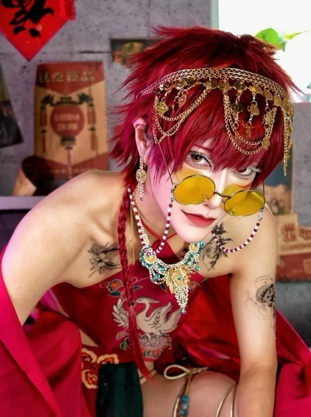 MIKECHI (みけち) with short red hair, round yellow glasses, gold chain headwear and layered jewelry, leaning toward the camera.