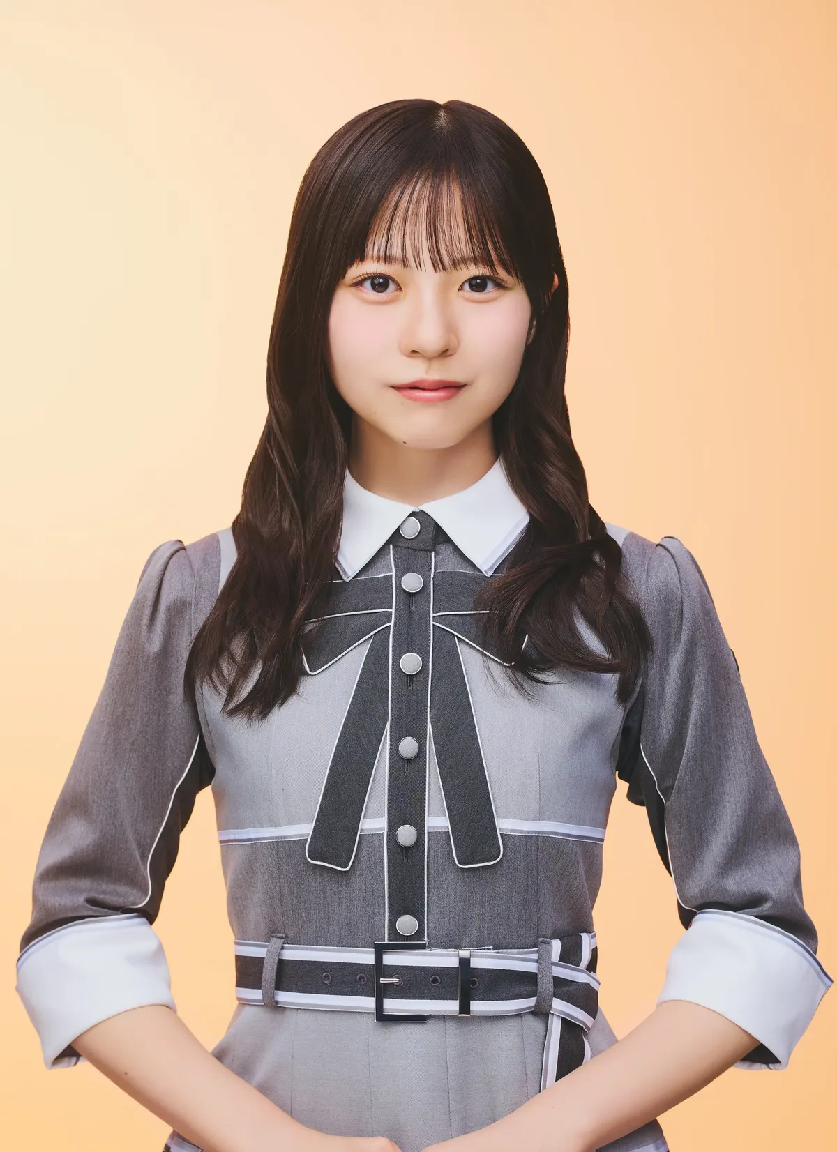 Yoko Shogenji faces the camera with her hands together, wearing a gray belted outfit with a white collar and dark ribbon against a pale orange background.
