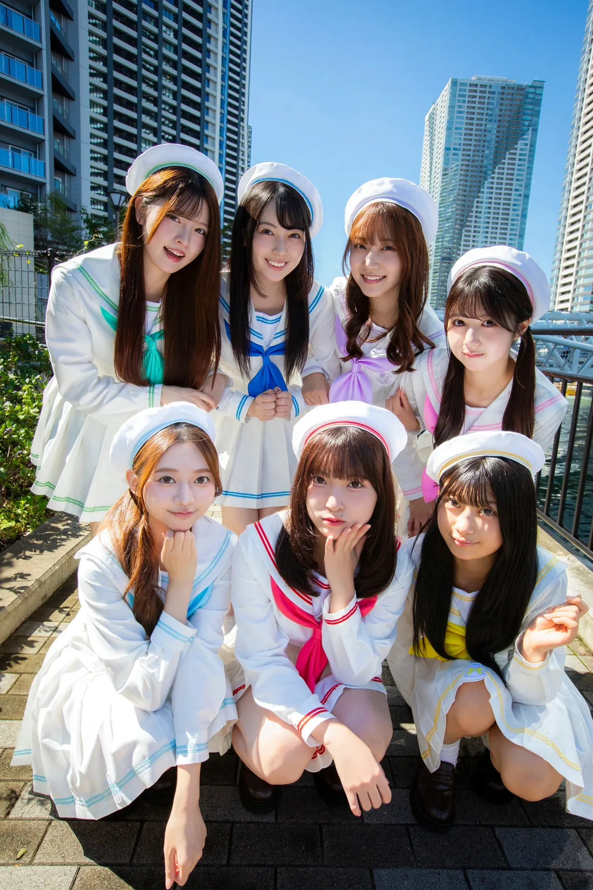 SUNNY PARADE’s seven members in white sailor-style outfits and hats, posing outdoors beneath a blue sky with high-rise buildings behind them.