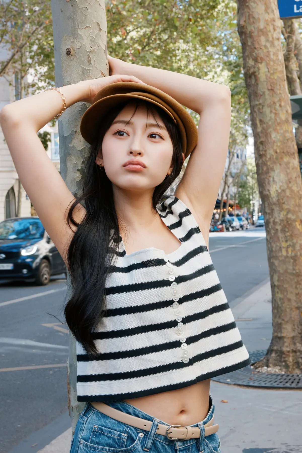 Kanami Takasaki in a brown cap, black-and-white striped sleeveless top and jeans, posing with her arms raised beside a street tree.